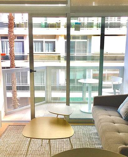 Salón luminoso con ventanal, sofá y mesas en un apartamento de Las Canteras diseñado por Interley.